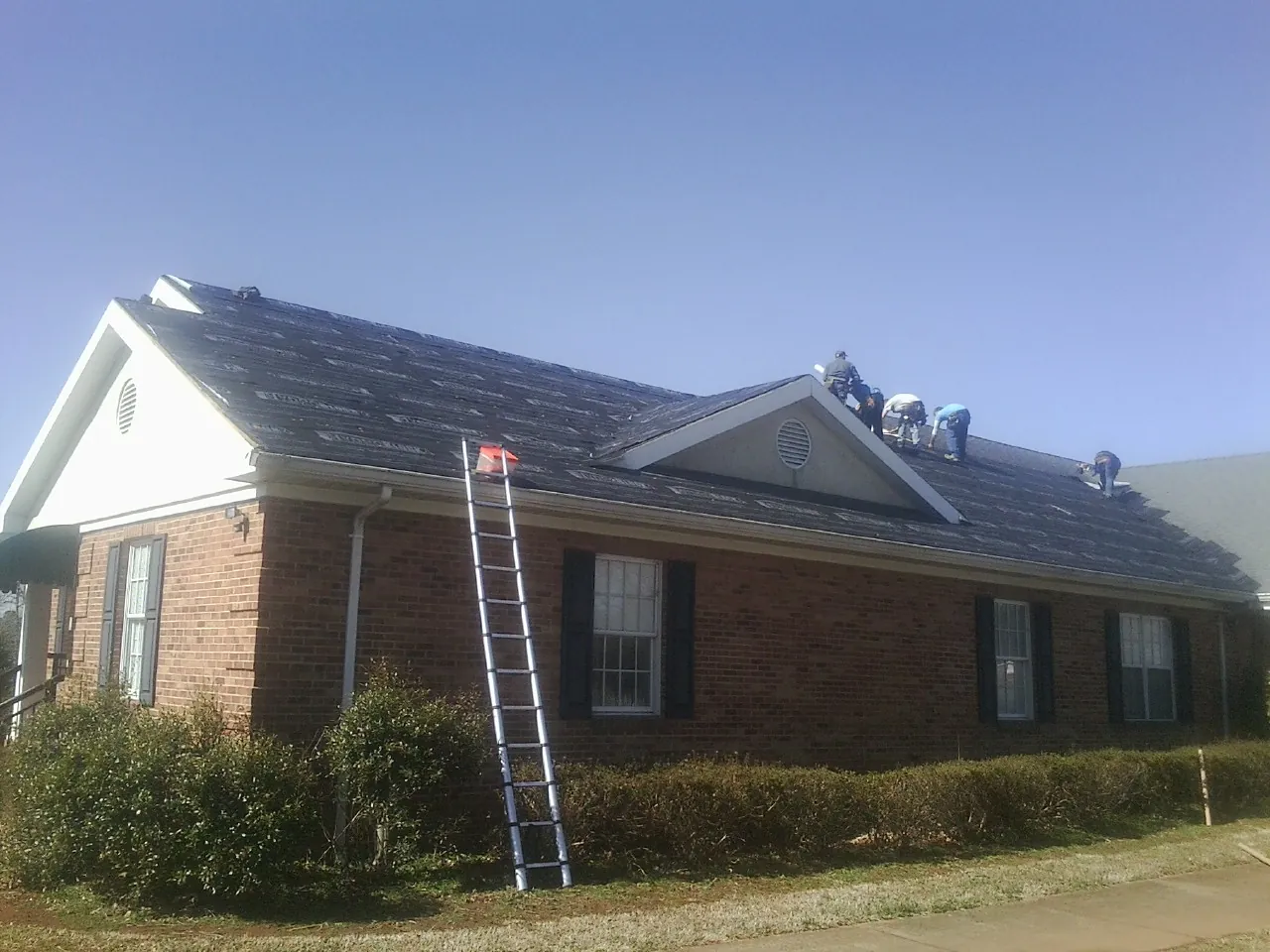 Crew tearing shingles off a brick ranch roof with a ladder set — Emergency Roof Repair work by our crew in New Ulm